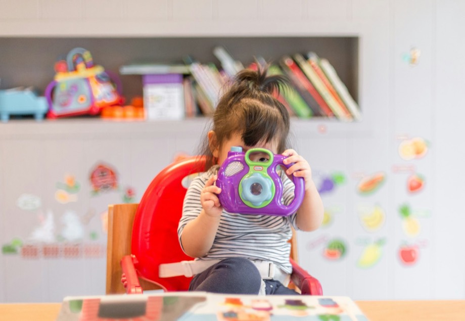A young child smiling and playing with a toy camera in a bright classroom